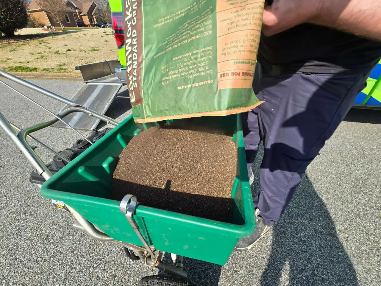 Lawn technician pouring organic granular fertilizer into a green broadcast spreader